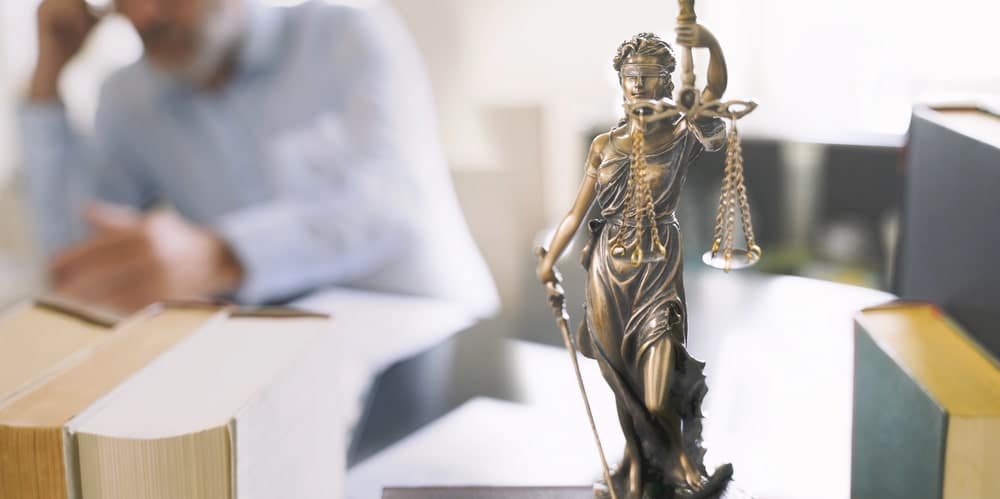 Image is of a Lady Justice statue in a law office with an attorney reviewing documents in the background, illustrating individual claim evaluation within a mass tort case in Nashville.