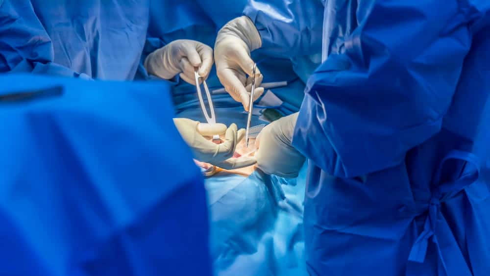 Image is of surgeons performing a procedure in an operating room, demonstrating the placement and use of surgical mesh in breast surgery.