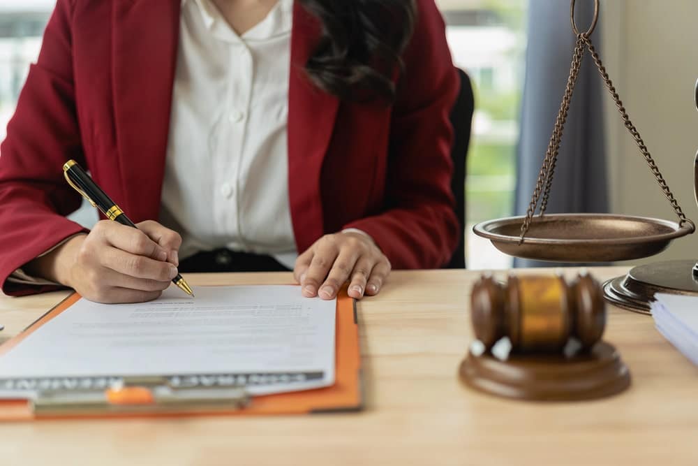 Image is of a person signing legal documents beside a judge's gavel and scales, concept of filing a claim involving defective breast mesh.