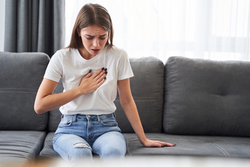 Image is of a woman experiencing chest pain while seated at home, concept of breast mesh injuries.