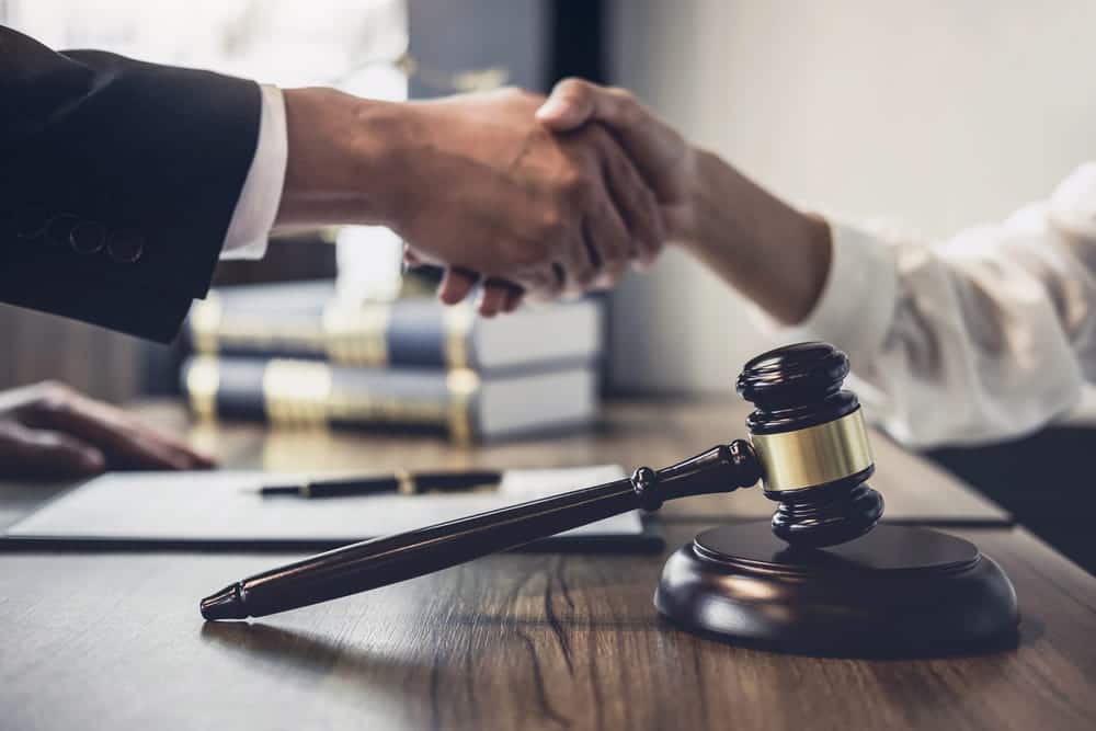 Image is of two people shaking hands beside a legal gavel, concept of settlement negotiation and reaching an agreement.